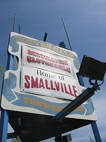 A white billboard with blue ribbon painted around the edges. In the ribbon, the words «Cloverdale Town Centre» appear in yellow surrounding two bannders. The top banner reads, «Destination Cloverdale», while the bottom reads «Home of Smallville».