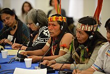 Cinq femmes sont installées à une tribune officielle équipée de micros. Parmi elles, deux portent des ornements remarquables comprenant des plumes et d'autres accessoires. On distingue d'autres personnes à l'arrière-plan et à l'avant-plan.