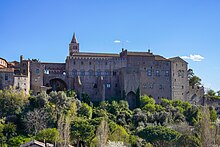 Um grande edifício, construído em tijolos, com uma torre, rodeada de árvores e pequenas casas