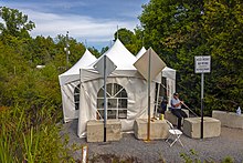 A group of small peaked-roofed white tents on a gravel road in a wooded area under a blue sky with clouds. In front of them are three cement blocks and three roads signs, two of which are diamond shaped with unfinished metal facing the camera, while the third, on the left, reads "Accés interdit aux piétons – No pedestrians". In front of the tent and on the right a man wearing a blue shirt and darker blue pants sits on a folding chair