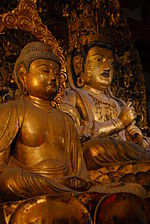 The central Bosatsu (in back) and one of four Great Bosatsu. Three-quarter view of two cross-legged seated statues (one in front of the other). The statue in the back appears larger and shows the Vajra Mudrā, enclosing the index finger of the left hand with the fist of the right hand. The statue in front has both hands resting on the lap.