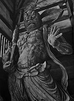 Agyō. Front view of a scary statue carrying a spear like object in his right hand and the palm of his left hand facing the viewer with fingers spread. Sculpted breastplate and necklace are visible.