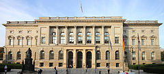 Frontale Farbfotografie eines hellen Sandsteingebäudes. Die Mitte des zweigeschossigen Gebäudes hat einen Vorbau mit Rundbögen und Säulen. Feine Ornamente verzieren die Fenstersimse und das Dach. Auf dem Vorplatz stehen zwei Bronzeskulpturen und drei Flaggen.