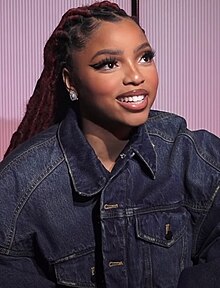 A woman with long, thick, dark hair behind her wears a blue denim jacket and smiles.