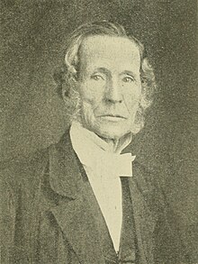 A sepia portrait of an elderly man with wavy side-parted hair, wearing a high-collared shirt with a cravat and a dark suit, gazing directly at the camera with a serious expression.