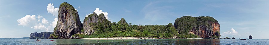 Em primeiro plano, o mar de águas azul, esverdeada. Ao centro, uma faixa de areia clara, com muitas pessoas. Alguns barcos podem ser vistos ao longe, próximo à praia. Além da praia, montanhas com paredões rochosos acinzentados a direita e à esquerda da praia, e montanhas mais suaves ao centro, cobertas pela floresta tropical.