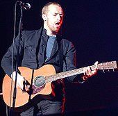 A man wearing a blue t-shirt and dark blue jacket holding a guitar and standing behind a microphone stand.