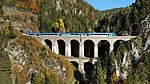 A photochorm picture of the a curving railroad bridge coming out of a tunnel bored into a large mountain. In the background another bridge and mountains are visible.