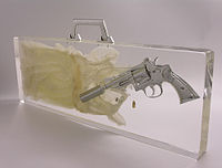 Ted Noten: Vrahova nevinnost, aktovka, stříbrný revolver, zlatá kulka, dětské křestní šaty, 2005, akrylátové sklo, sbírka Stedelijk Museum. Fotografováno při horním šikmém protisvětle na nekonečném bílém pozadí.