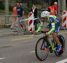 A cyclist riding a bicycle with a solid rear wheel and wearing a skin-tight lime green and blue jersey with white trim, with an aerodynamic helmet. Spectators watch him from the roadside.