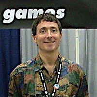 A man wearing multicolored shirt smiles at the camera. Behind him stands a blue and white background, above which is an excerpt of logo which reads "games".