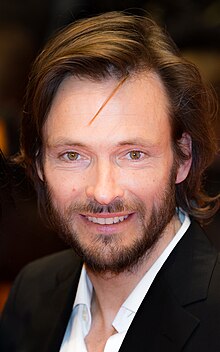 close-up of Andreas Pietschmann wearing a white shirt and black blazer, grinning at camera