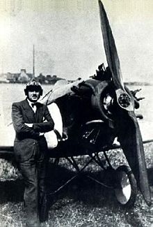 A black and white photo of a man in a suit and bow tie, wearing a pilot helmet, standing in front of a small propeller-driven monoplane