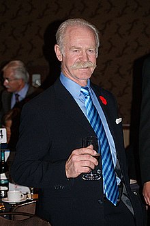 A grey-haired, grey-moustached man holds a wine glass. He is wearing a blue suit with a striped blue tie