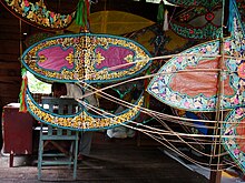 Man in a workshop making Waus; a purple wau with gold outlines hangs in front of him, as does an empty frame