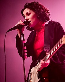 A woman performs at a concert; she is holding a guitar in one hand and a microphone in the other