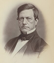 Black and white photograph of a Caucasian male with a combed-over dark hair wearing a bowtie, a black suit, and a starched white collar