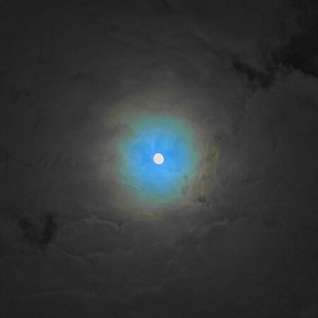 Der Mond im Goldenen Tor der Ekliptik bei leichter Bewölkung mit mehrfarbiger Korona (unten im Bild der Rote Riese Aldebaran, oben rechts die Plejaden). Die Farbe der Wolken ist im neutralen Grau (Farbtemperatur des Mondlichts = 4100 Kelvin).