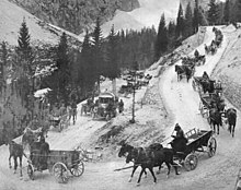 Le long d'un virage de lacets, au premier plan des caravanes de ravitaillement se succèdent, à l'arrière plan des falaises de montagnes italiennes.