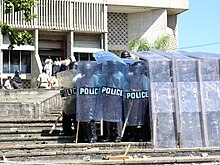 Plusieurs policiers armés en uniforme noir avec casques et boucliers. Protégés par un écran transparent en pvc sur un escalier devant un édifice.