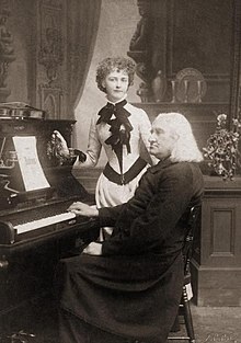 Arma Senkrah standing next to Franz Liszt who is sitting at a piano