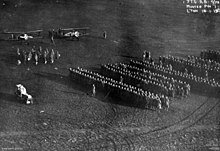 A large number of people standing in formation in a field. Two biplanes are visible at the upper left, with a smaller group of people standing in front of them.