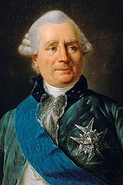 Middle-aged, white-haired man wearing a blue velvet jacket, white shirt, and a big piece of jewelry pinned to his jacket.