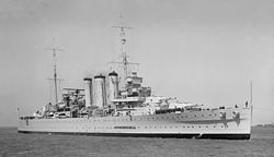 Photograph of a war ship in a body of water. Large cannons can be seen on the deck, along with sailors.