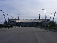 A straight tarmac road. At the head of the road is a stadium with a bowl-shaped outline, surrounded by a number of masts, with cables running to the stadium roof