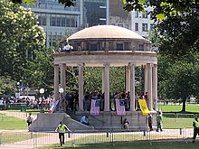 A group of people stand in a bandstand in a park, displaying various American flags and the Gadsden Flag. They are surrounded by metal barricades protected by police.