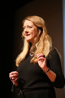 Annie Machon standing, left hand raised, speaking into a headset microphone