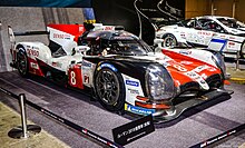 Side view of a red, white and black Toyota TS050 Hybrid enclosed in a space at an automobile show