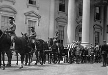 A black-and-white photograph of a procession of people in uniforms, some of whom are riding horses, all standing in front of a white building