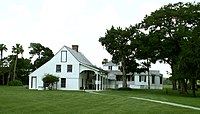 A color photo of a white two-story building with black shutters, a tall wood-shingled roof, and an outdoor veranda. The first floor has a stucco finish and has one window, the second story is wood clapboard and has two windows topped with a chimney. A lawn and trees surround it.