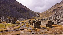 Photographie couleur des vestiges d'un village de mineurs près de Glendalough avec un ruisseau serpentant entre les anciennes bâtisses et des éboulis, avec une végétation très pauvre