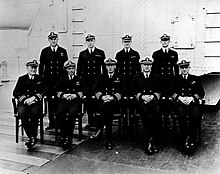 Two rows of uniformed naval officers, five seated and four standing, in a black and white photograph taken on a ship's deck