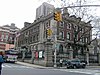 A three-story brick and stone building sits on a corner in New York City and is surrounded by a stone and wrought iron fence.