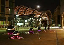 Millennium Square (adjacent to the Peace Gardens) and Sheffield Winter Gardens, at night. Vsisble are several large, lit, steel balls at the centre of the square and the modern architecture of the Winter Gardens is visible in the background.