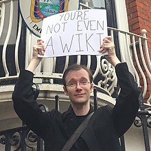 Garrett in 2016, standing outside the Ecuadorian Embassy in London, where WikiLeaks founder Julian Assange had received asylum, holding a sign saying "You're not even a Wiki"