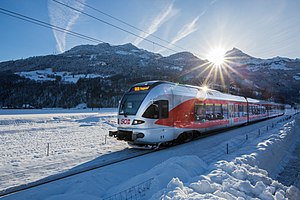 Silver and red train next to racing past a snow-covered field with mountains in the background