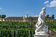 Farbfotografie in der Untersicht mehrerer Weinbergterrassen mit einer Treppe in deren Mitte. Rechts unten eine weiße Frauenfigur auf einem Sockel. Im Hintergrund ist ein gelbes Schloss mit vielen Rundbogenfenstern und dunklen Steinskulpturen dazwischen. Auf dem runden Mittelteil ist eine grüne Kuppel.