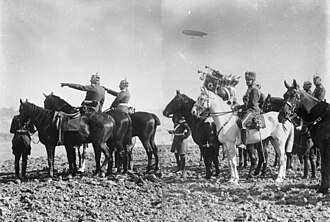 Photo de la suite impériale à cheval, avec les trompettes des garde du corps en train de sonner, le tout surmonté par un dirigeable.