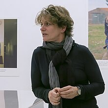 Bettina Flitner ist mittig stehend in Galerieräumen zu sehen. Sie steht nach rechts gewandt im Gespräch, die Hände auf Höhe der Körpermitte haltend. Sie trägt einen schwarzen langärmligen Pullover. Um den Hals trägt sie einen Schal, der abwechselnd, schwarz, grau und dunkelgrün und mehrfach um ihren Hals gebunden ist und dessen Ende aus einem Knoten raushängt. Sie hat kurzes dunkelblondes, zerzaustes Haar.