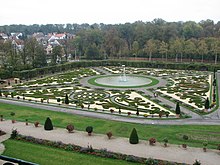 Farbfotografie in der Obersicht eines Springbrunnens, der von einem ornamentalen Beet auf hellem Boden umkreist ist. An den Außenrändern der Beete stehen Blumentöpfe mit kleinen Bäumen und Blumensträuchern. Im Vordergrund stehen weitere Blumentöpfe auf einem Weg mit schmaler Rasenfläche dahinter. Im Hintergrund sind links ein Ort und rechts Wald zu sehen.