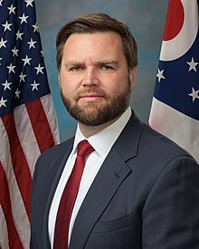Vance poses for a professional portrait in a suit and red tie. Behind him the flag of the US is partly visible on his left and the flag of Ohio on the right.