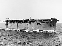 Black and white photograph of an aircraft carrier at sea. An aircraft is about to land on her stern, and another aircraft is positioned at the forward end of the flight deck