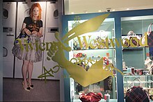 Blick durch ein Schaufenster mit goldenem Aufkleber, der das Logo und einen Säbel schwingenden Arm beinhaltet. Im Laden ist links ein Fotoposter mit Westwood in einem Waschsalon und rechts eine blaue Vitrine mit Accessoires und Handtaschen zu sehen.