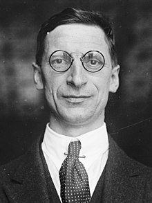 Portrait en noir et blanc d’un homme en costume, portant des binocles, qui fixe l’objectif avec un léger sourire.