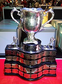 Silver bowl trophy with two large handles, mounted on a wide black plinth engraved with team names on silver plates.