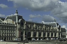 An ornate building, with peaked roofs at either end and large clocks on their fronts, under a blue sky with clouds, seen from its front right